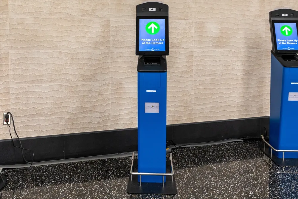 A blue Global Entry kiosk with an upright display reading Please Look Up at the Camera next to a neutral textured wall.