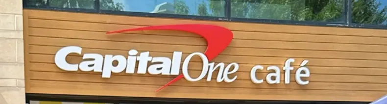 Exterior signage for Capital One Café featuring white raised lettering and a red swoosh logo on horizontal wood paneling.