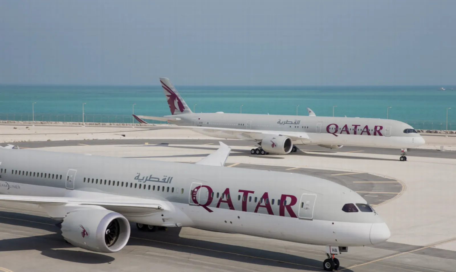 Two Qatar Airways passenger jets parked on a coastal airport apron beside turquoise water.