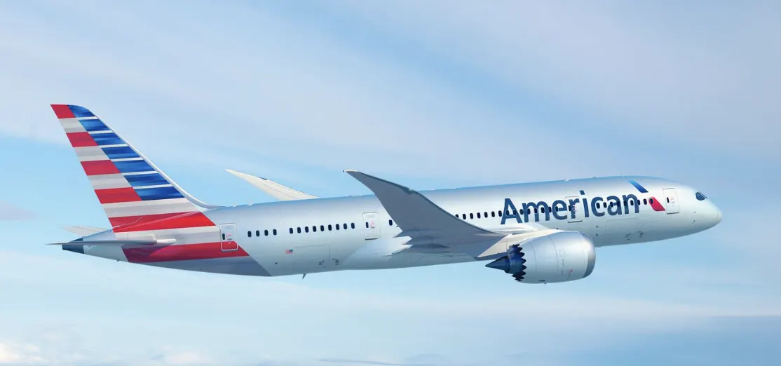 An American Airlines passenger jet flies through a bright blue sky with faint white clouds.